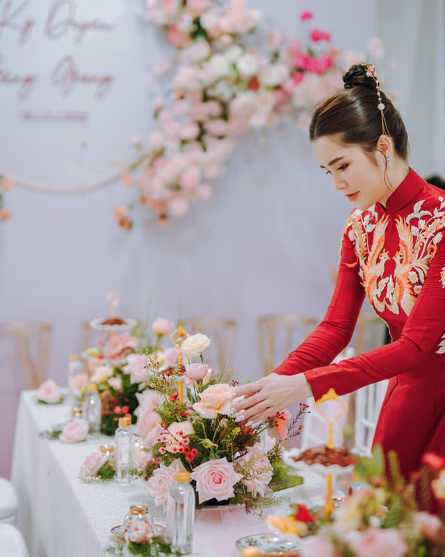 An Asian woman in traditional red dress arranges a floral centerpiece at an elegant indoor event.