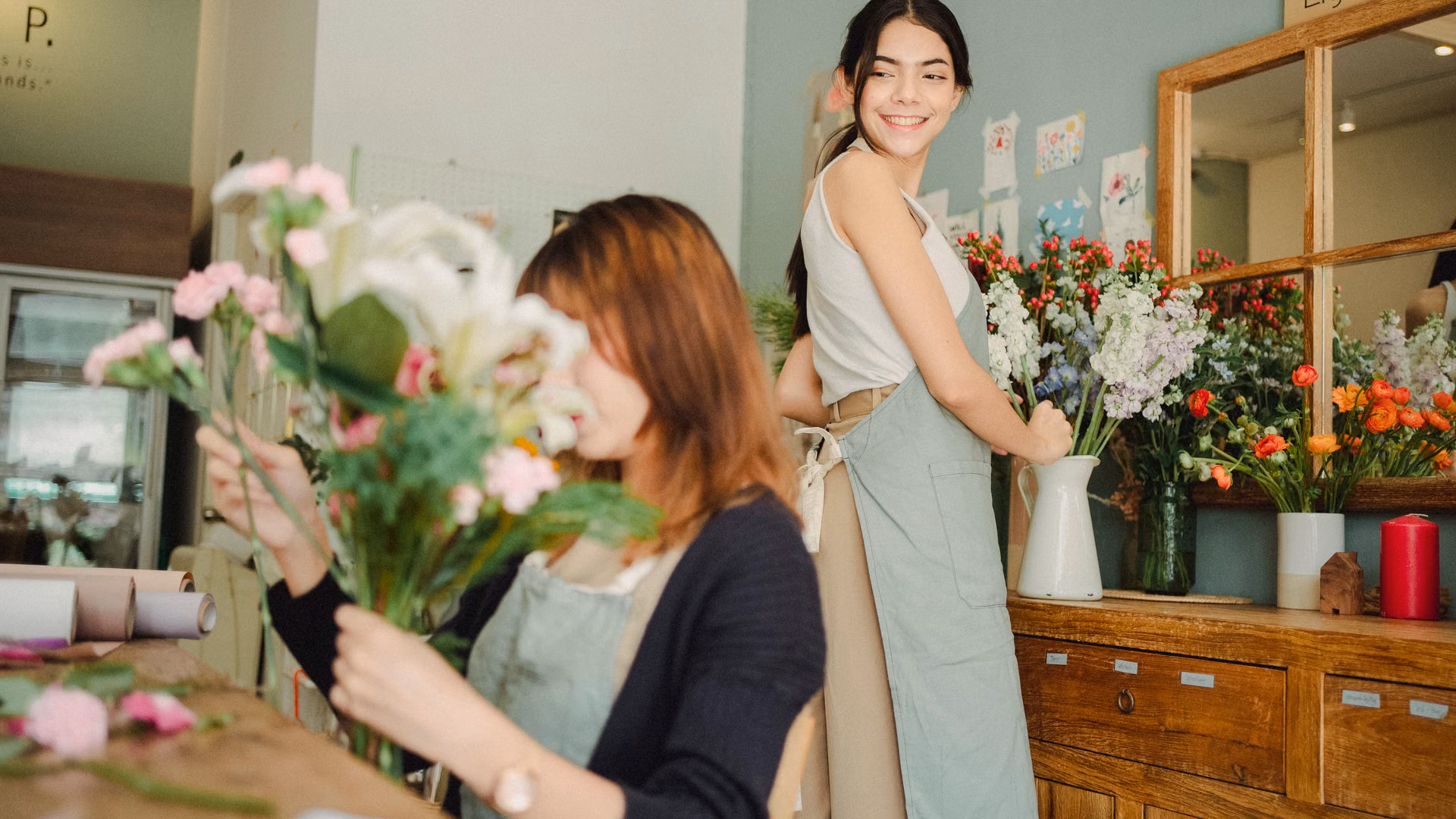 Side view of smiling female florist wearing apron making bouquet of blooming flowers while working with coworker in floral workshop