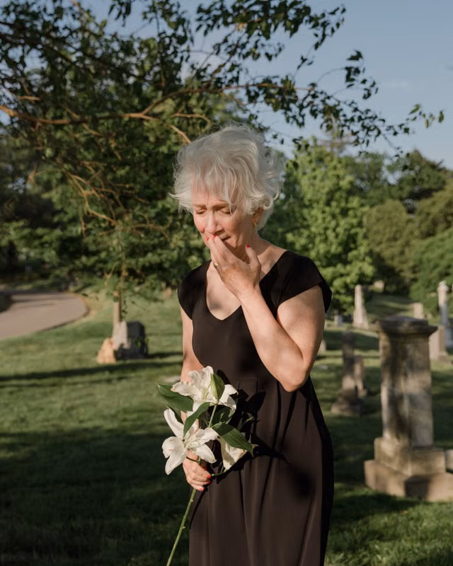 An elderly woman stands in a cemetery, holding white lilies and expressing grief.